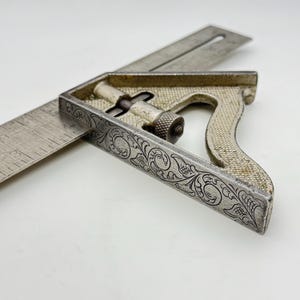 May include: A vintage combination square with a silver finish. The tool features a ruler and a handle with ornate floral engravings. The square is designed for measuring and marking angles, and is a classic woodworking tool.