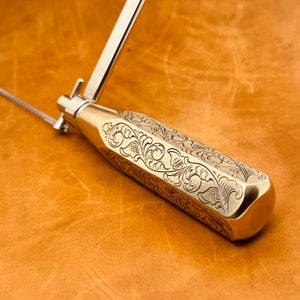 May include: A close-up of a brass saw with a decorative, engraved handle. The handle features a floral design with intricate details.