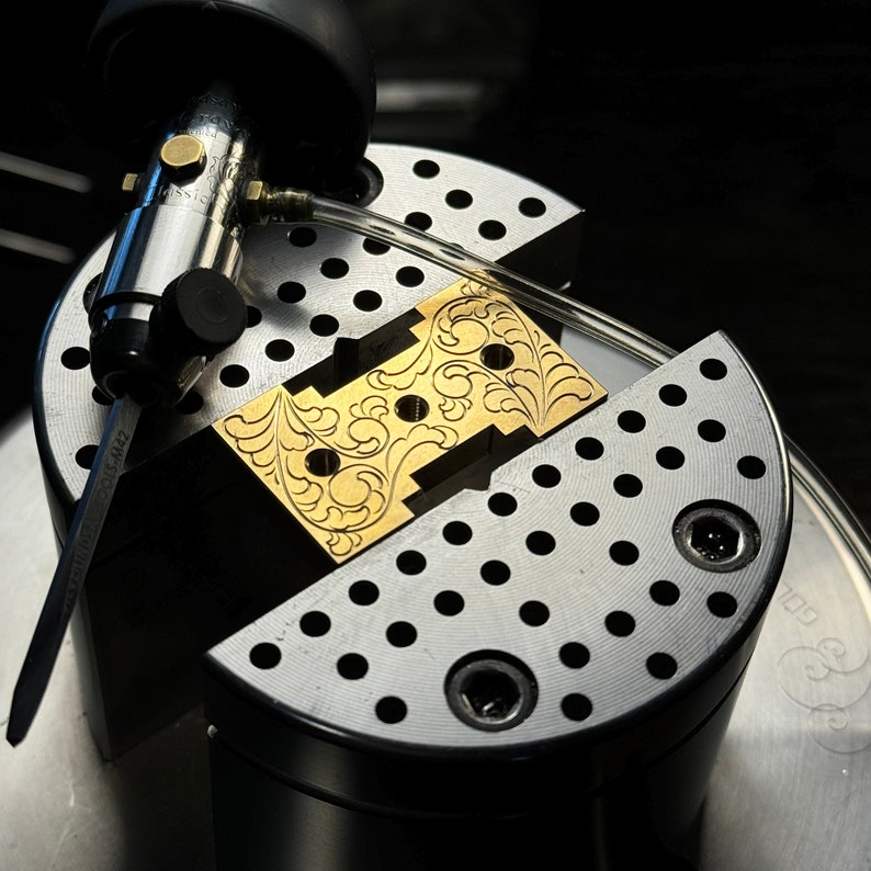 May include: A gold-colored metal piece with an intricate floral design is clamped in a vise. The vise is made of metal and has a grid pattern.