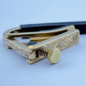 May include: A gold-colored guitar capo with an intricate floral design. The capo has a black rubber pad and a spring-loaded lever.