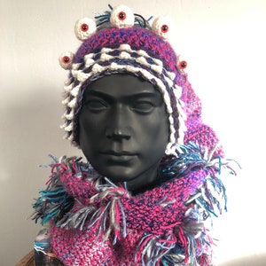 Puede incluir: Un gorro de crochet rosa, azul y blanco con flecos. El gorro tiene una fila de ojos blancos de crochet con pupilas rojas alrededor de la parte superior.