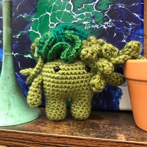 Könnte beinhalten: Ein grünes Amigurumi-Spielzeug aus Häkelarbeit mit einem Blatt-Kopf und einem freundlichen Gesicht. Das Spielzeug sitzt auf einer Holzoberfläche.