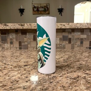 Puede incluir: Vaso cil&iacute;ndrico blanco con el logotipo verde de Starbucks y un dise&ntilde;o de Grogu. El vaso est&aacute; sobre una encimera moteada. El dise&ntilde;o incorpora el logotipo de Starbucks y un personaje de The Mandalorian.