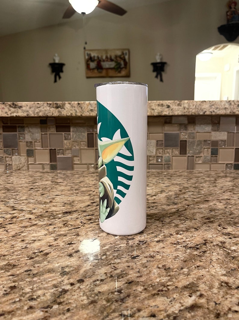 Puede incluir: Vaso cil&iacute;ndrico blanco con el logotipo verde de Starbucks y un dise&ntilde;o de Grogu. El vaso est&aacute; sobre una encimera moteada. El dise&ntilde;o incorpora el logotipo de Starbucks y un personaje de The Mandalorian.