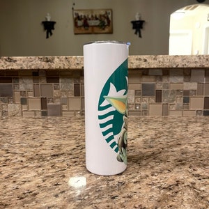 Puede incluir: Un vaso blanco con un dise&ntilde;o verde y blanco de un personaje de dibujos animados. El vaso tiene una tapa plateada y una forma cil&iacute;ndrica y delgada. El dise&ntilde;o incorpora un logotipo estilizado con la imagen del personaje.