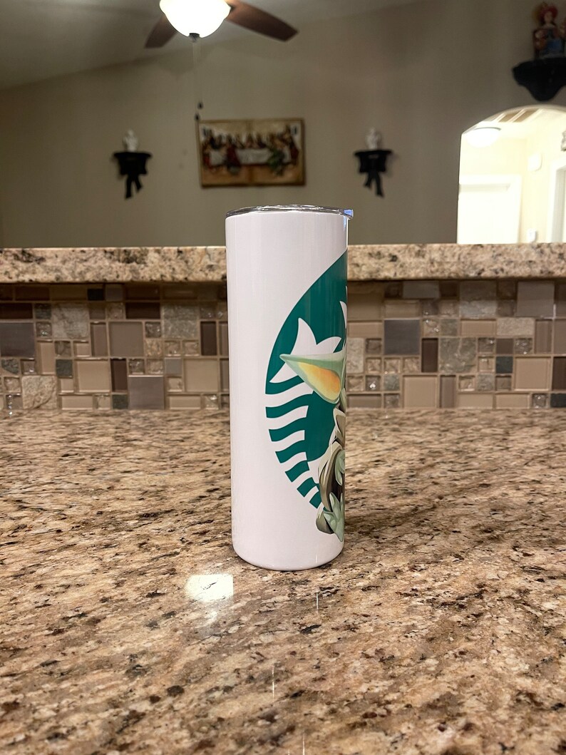 Puede incluir: Un vaso blanco con un dise&ntilde;o verde y blanco de un personaje de dibujos animados. El vaso tiene una tapa plateada y una forma cil&iacute;ndrica y delgada. El dise&ntilde;o incorpora un logotipo estilizado con la imagen del personaje.
