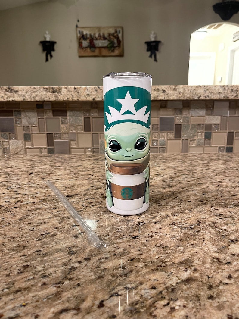 Puede incluir: Un vaso de acero inoxidable con el logotipo verde y blanco de Starbucks y una imagen de dibujos animados de Grogu de The Mandalorian. El vaso tambi&eacute;n tiene una pajita de pl&aacute;stico transparente.