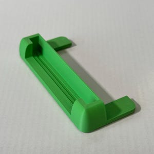 May include: A bright green 3D printed plastic bracket featuring a long horizontal channel with raised side supports and two flat mounting tabs extending from the back. The object has a smooth finish with visible layer lines from the printing process.