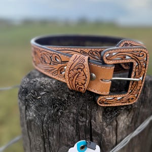 May include: A light brown leather belt with detailed floral and scrollwork carving. The belt has a decorative buckle and a dark blue interior. The belt is displayed on a wooden post with a tag attached.