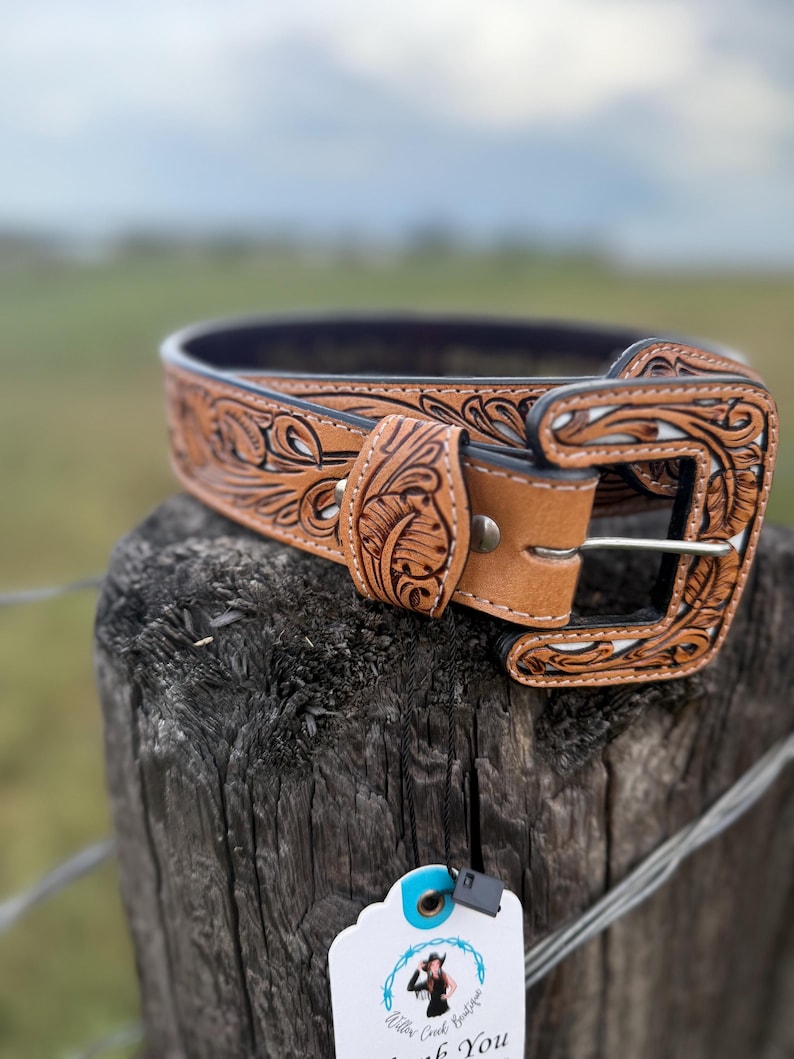 May include: A light brown leather belt with detailed floral and scrollwork carving. The belt has a decorative buckle and a dark blue interior. The belt is displayed on a wooden post with a tag attached.