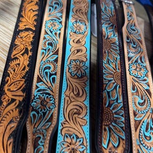May include: Close-up of several tooled leather straps with floral and sunflower designs. The straps feature brown, black, and turquoise colors. The intricate patterns are embossed on the leather, showcasing a Western or country aesthetic.