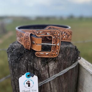 May include: A brown leather belt with detailed floral and scroll patterns. The belt has a silver buckle and a white background on the buckle. A tag with the words "Thank You" is visible below the belt.