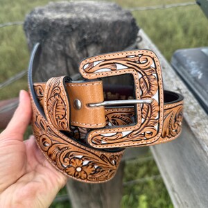 May include: A brown leather belt with detailed floral and leaf patterns. The belt has a light brown buckle and keeper, with white detailing in the carved design. The belt is displayed against a natural outdoor backdrop.