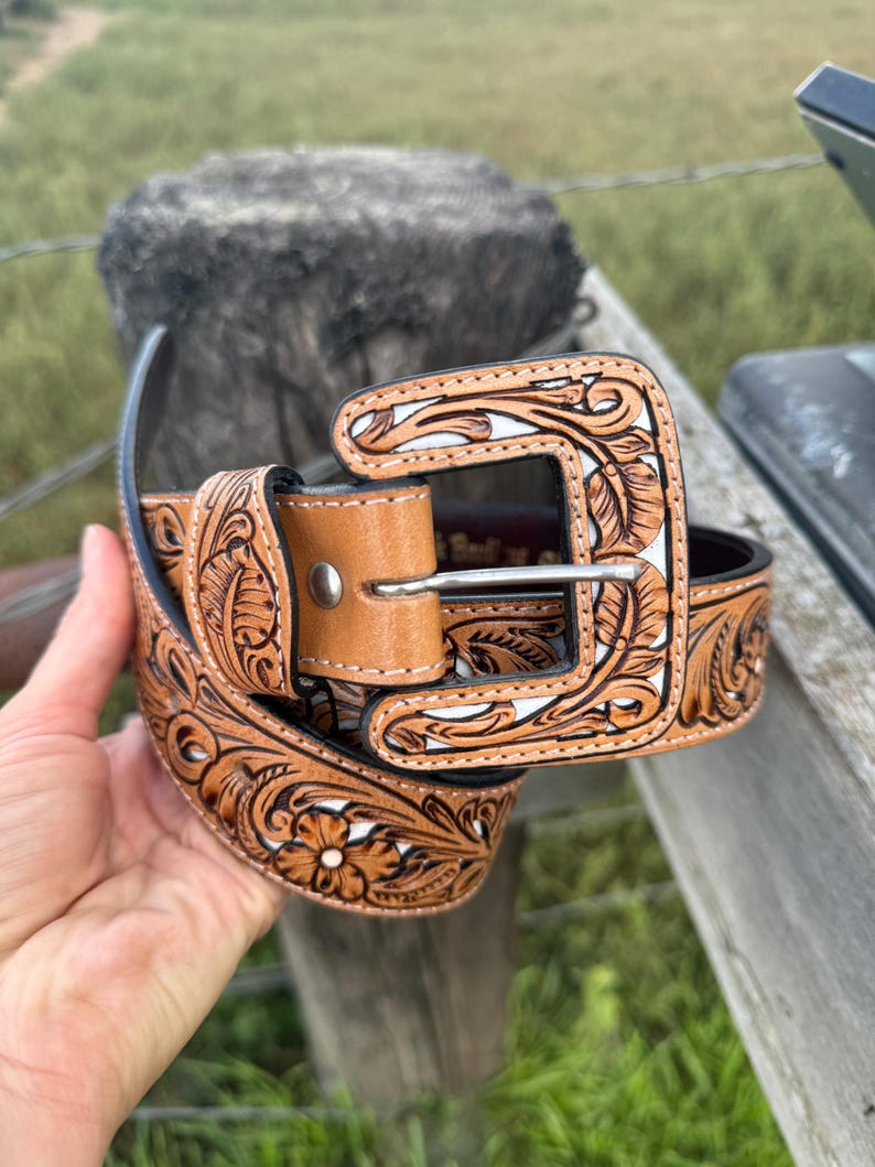 May include: A brown leather belt with detailed floral and leaf patterns. The belt has a light brown buckle and keeper, with white detailing in the carved design. The belt is displayed against a natural outdoor backdrop.