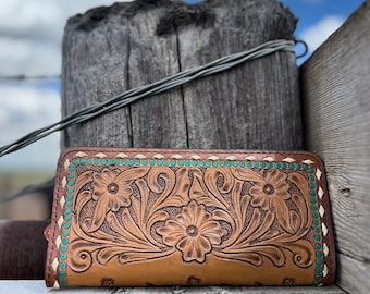 Puede incluir: Una cartera de cuero marrón con un diseño floral y un ribete turquesa y blanco. La cartera está sobre una superficie de madera.