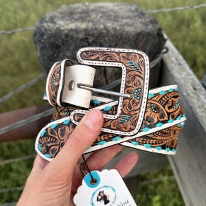 May include: A brown leather belt with floral tooling and turquoise details. The belt has a white buckle and keeper. A tag with "Thank You" is attached. The belt is approximately 3.8 cm wide.