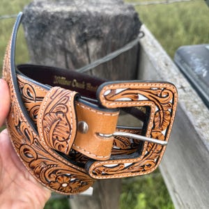 May include: A brown leather belt with detailed floral and leaf patterns. The belt has a light brown buckle and a silver-coloured metal buckle. The text "Willow Creek Belts" is visible on the belt.