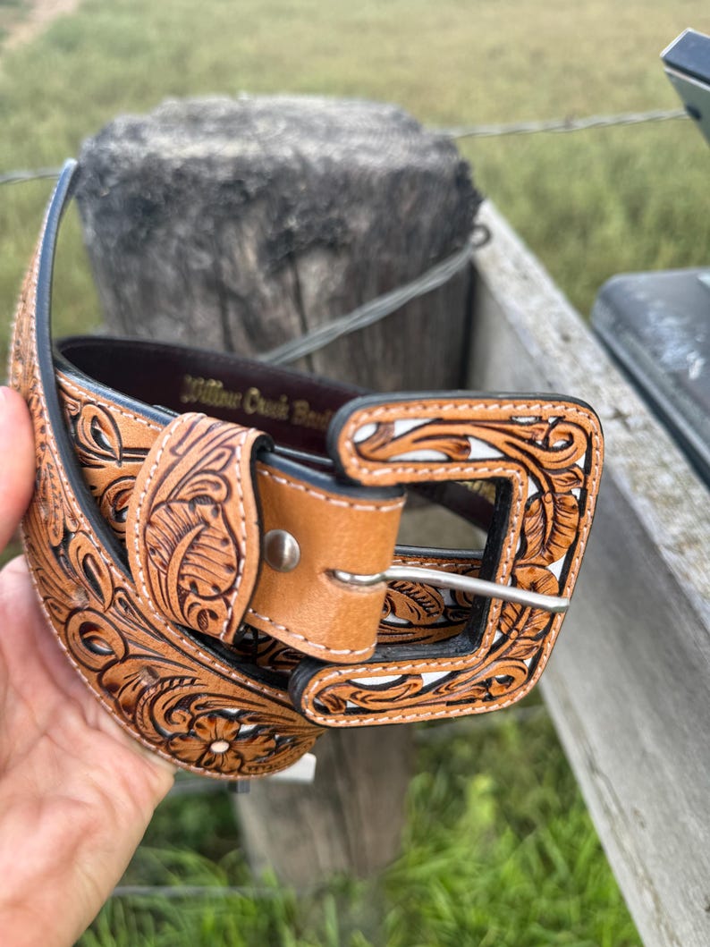 May include: A brown leather belt with detailed floral and leaf patterns. The belt has a light brown buckle and a silver-coloured metal buckle. The text "Willow Creek Belts" is visible on the belt.