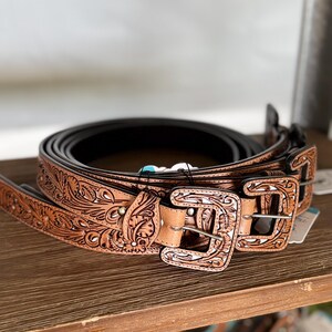 May include: A collection of brown leather belts with detailed floral and scroll patterns. Each belt has a decorative buckle. The belts are stacked on a wooden shelf, highlighting the craftsmanship and design of the belts.