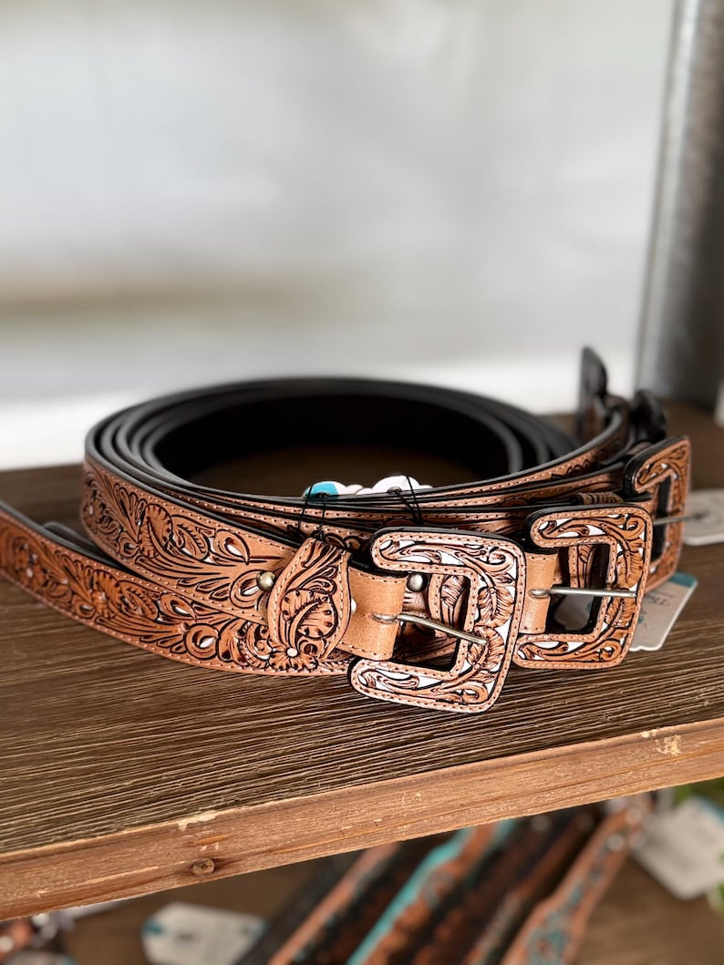 May include: A collection of brown leather belts with detailed floral and scroll patterns. Each belt has a decorative buckle. The belts are stacked on a wooden shelf, highlighting the craftsmanship and design of the belts.