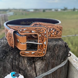May include: A tan leather belt with a floral pattern and a silver buckle. The belt features carved floral designs with a white background. The belt is displayed on a wooden post with a tag that says "Thank You".