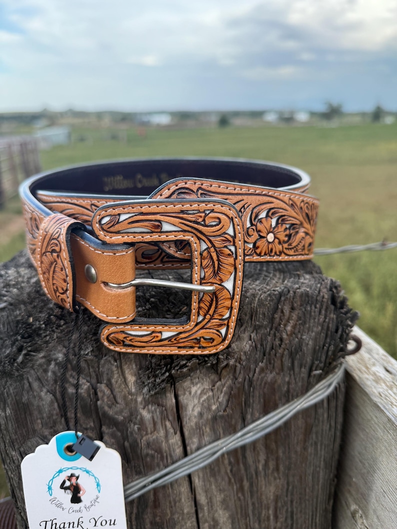 May include: A tan leather belt with a floral pattern and a silver buckle. The belt features carved floral designs with a white background. The belt is displayed on a wooden post with a tag that says "Thank You".
