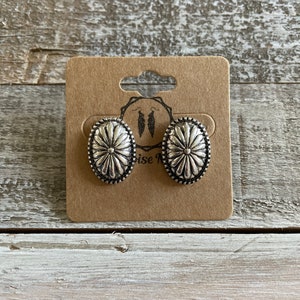 May include: A pair of silver-toned oval stud earrings with a floral design, displayed on a brown card. The earrings have a detailed, textured surface and a dark border. The card has a cutout at the top and a logo with feather illustrations.