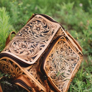 May include: A tan leather backpack featuring intricate hand-tooled floral patterns with dark brown accents. The bag includes a large main compartment and a smaller front pocket, both secured with zippers, showcasing detailed carved leaf and flower motifs.