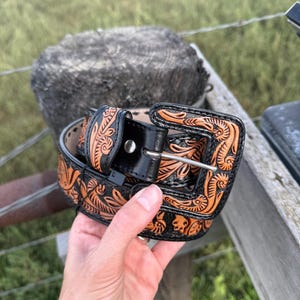 May include: A black leather belt with detailed floral and scrollwork patterns in tan. The belt has a silver-coloured buckle. The belt is held by a hand against a blurred outdoor backdrop. The belt is a western-style accessory, suitable for waist sizes.