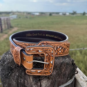 May include: A brown leather belt with a floral tooled design and a silver buckle. The belt is displayed on a wooden post with a blurred background of a field and sky. The belt has the text "Willow Creek Ranch" and "Genuine Leather".