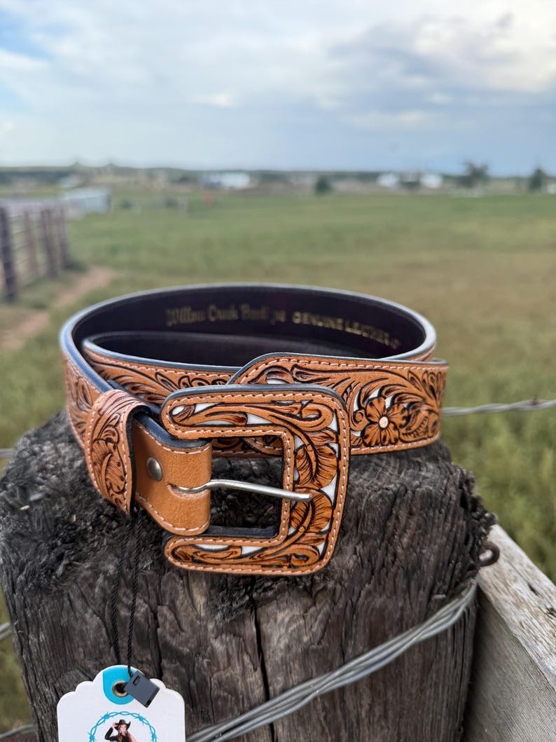 May include: A brown leather belt with a floral tooled design and a silver buckle. The belt is displayed on a wooden post with a blurred background of a field and sky. The belt has the text "Willow Creek Ranch" and "Genuine Leather".