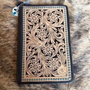 May include: Black leather journal with a tooled floral design in tan and turquoise accents. The cover features a detailed pattern of flowers and leaves, with a turquoise border and a small emblem in the corner. The journal is closed with a zipper.