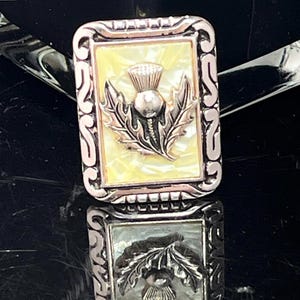 May include: Rectangular silver-toned brooch with a thistle design. The brooch features a light yellow background with a silver thistle and leaves. The frame has a decorative, ornate design. The brooch is reflected in the surface below.