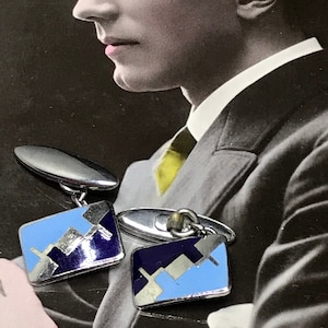 May include: A pair of silver cufflinks with blue and white enamel geometric designs. The cufflinks are in the shape of a rectangle with rounded corners.