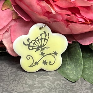 Puede incluir: Un amuleto de cerámica amarillo con forma de flor con un diseño de mariposa negra y un lazo plateado para colgar.