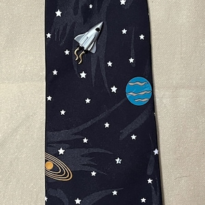 May include: A black tie with a space theme. It features white stars, a crescent moon, a rocket ship, a red planet with rings, a blue planet with rings, and a green and blue planet.