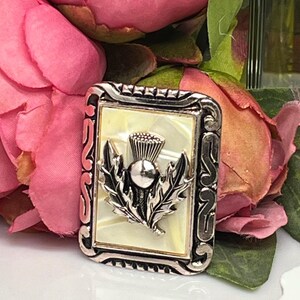 Puede incluir: Un broche plateado con un fondo de nácar blanco con un diseño de cardo plateado.