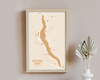May include: A beige and brown illustrated map of Cayuga Lake, New York, with the text "Cayuga Lake Wineries" printed below the map. The map is framed in a light wood frame and is hanging on a light beige wall.