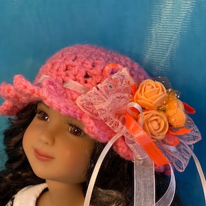 May include: A pink crocheted hat with a white lace trim and orange fabric flowers. The hat is decorated with white and orange ribbons.