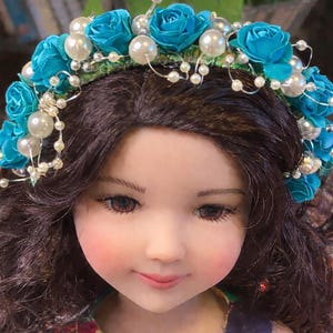 Puede incluir: Una muñeca con cabello castaño y una corona floral turquesa con detalles de perlas. La muñeca tiene mejillas rosadas y viste un vestido con estampado floral. La corona presenta rosas de tela turquesa y pequeñas perlas blancas.