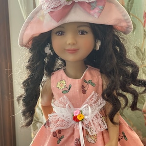 May include: A doll wearing a pink dress with a white lace bow and a pink and white hat with a white lace bow. The doll has brown hair and brown eyes.
