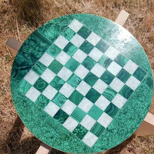 Può includere: Un tavolo da scacchi rotondo con una superficie verde malachite e un intarsio a scacchiera. La scacchiera presenta quadrati verdi e bianchi alternati. Il tavolo è sostenuto da quattro gambe in legno, creando un pezzo unico e decorativo.