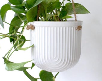 以下が含まれることがあります： 緑の植物が入った2つの白いハンギングプランター。プランターはリブのデザインで、茶色のロープで吊り下げられています。片方のプランターはもう一方より大きいです。植物は緑の葉を持っています。