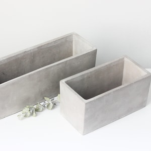 May include: Two rectangular, gray concrete planters of different sizes. The larger planter is positioned behind the smaller one. Both planters have a simple, modern design, suitable for indoor or outdoor use. A few sprigs of greenery are placed between the planters.