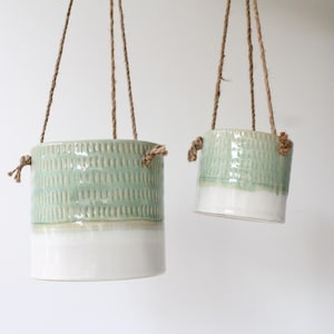 Peut inclure: Deux pots de fleurs suspendus en céramique. Les pots sont cylindriques avec une partie supérieure texturée vert clair et une partie inférieure blanche. Ils sont suspendus par une corde en fibre naturelle.
