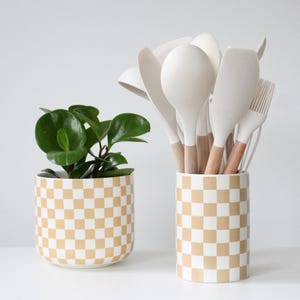 May include: A small potted plant with green leaves sits next to a ceramic utensil holder. The holder and pot have a tan and white checkered pattern. The holder contains white cooking utensils with wooden handles.