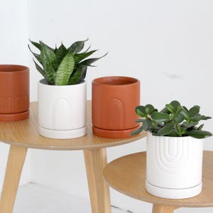 May include: Four ceramic planters in shades of terracotta and white, each with a matching saucer. Two planters hold green plants. The planters are on two small, round wooden tables with three legs.