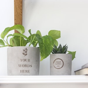 Peut inclure: Deux pots de fleurs en béton gris avec des plantes vertes. Le plus grand pot porte l'inscription "YOUR WORDS HERE" et un motif botanique. Le plus petit pot porte l'inscription "YOUR LOGO HERE" dans un cercle. Des livres sont sur une étagère.