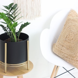 May include: A black planter with gold-colored legs and trim holds a green plant. A beige woven pillow rests on a white chair. A macrame wall hanging is in the background. The scene is bright and airy.