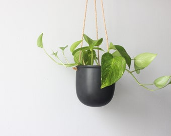 May include: A matte black, rounded hanging planter suspended by three thin, light brown jute ropes. A trailing pothos plant with variegated green and yellow heart-shaped leaves grows out of the container, with vines extending outward to the sides.